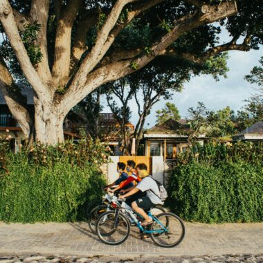 Three boys ride bicycles along a cobblestone street bordered by lush greenery and traditional houses in Bali.