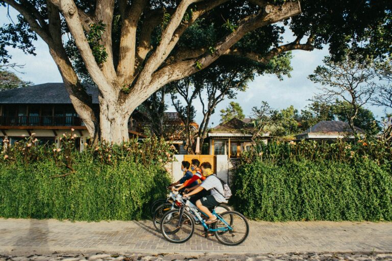 Three boys ride bicycles along a cobblestone street bordered by lush greenery and traditional houses in Bali.